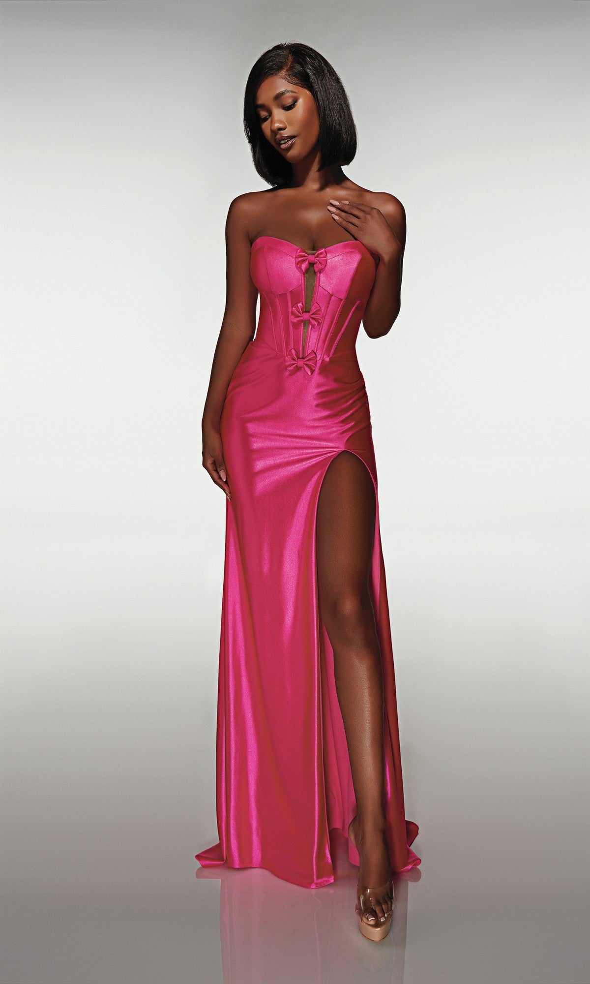 A woman in the Alyce 62046 long prom dress—strapless, metallic, with a corset bodice and thigh-high slit—stands against a plain light background wearing clear high heels.
